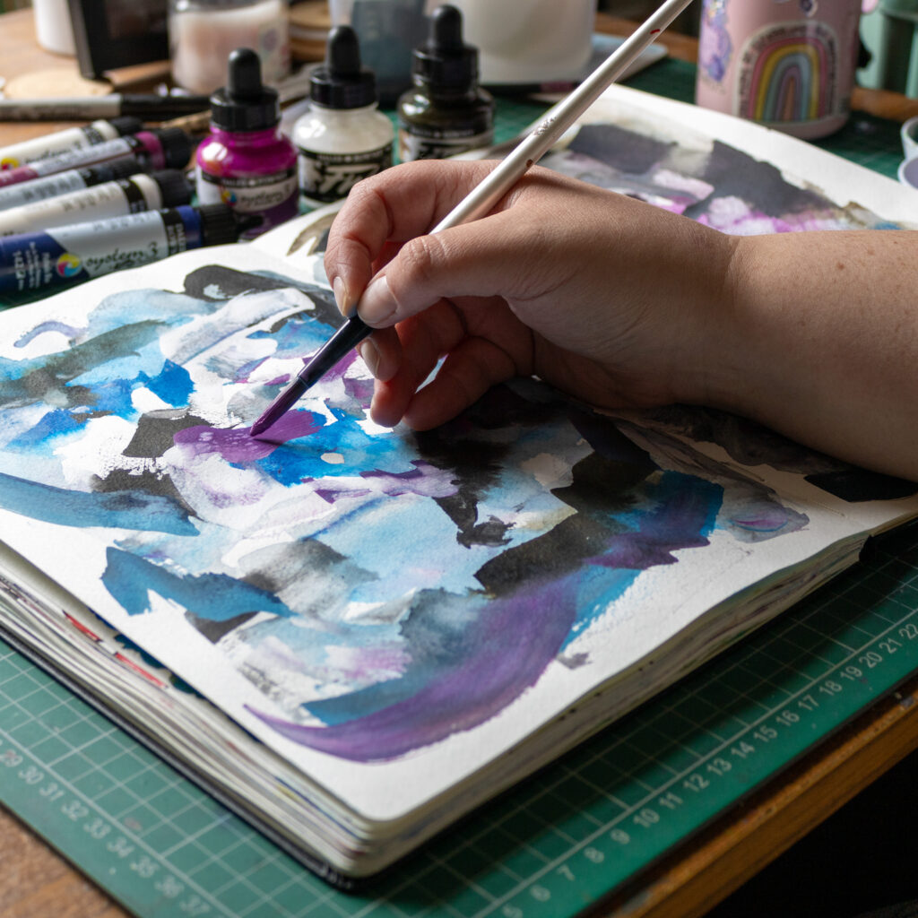 A photo of an open sketchbook sat on top of a green cutting mat and surrounded by a range of ink bottles in pinks and blues. A white person is holding a paint brush and is making abstract marks on the page.